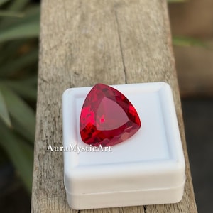 Puede incluir: Una gema roja oscura, tallada en forma triangular, descansa en una caja blanca. La gema está facetada y es brillante. El texto "AuraMysticArt" está impreso en la caja. El fondo es de madera.