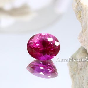 May include: A round, faceted, hot pink gemstone with a brilliant cut. The gem is reflected on a mirrored surface. The text "AuraMysticA" is visible. The gem is set against a white background with a piece of white rock.