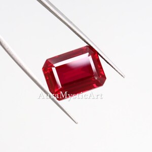 May include: A vibrant, rectangular-cut red gemstone held by silver-toned metal tweezers. The stone features a faceted surface with deep crimson hues and bright light reflections. The text AuraMysticArt is visible across the centre of the stone.