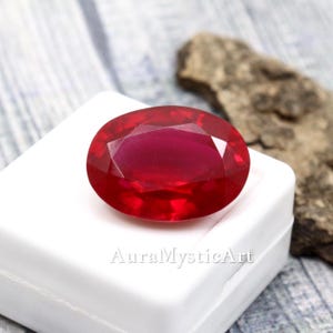May include: A deep red, oval-cut ruby gemstone, set on a white surface. The image shows the text "AuraMysticArt". The background features a wooden texture and a piece of natural stone.