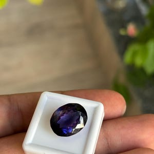 May include: A faceted, oval-shaped, deep purple gemstone set in a white square display case. The gemstone has a brilliant cut, reflecting light. The background is blurred, with hints of green foliage and a wooden surface.