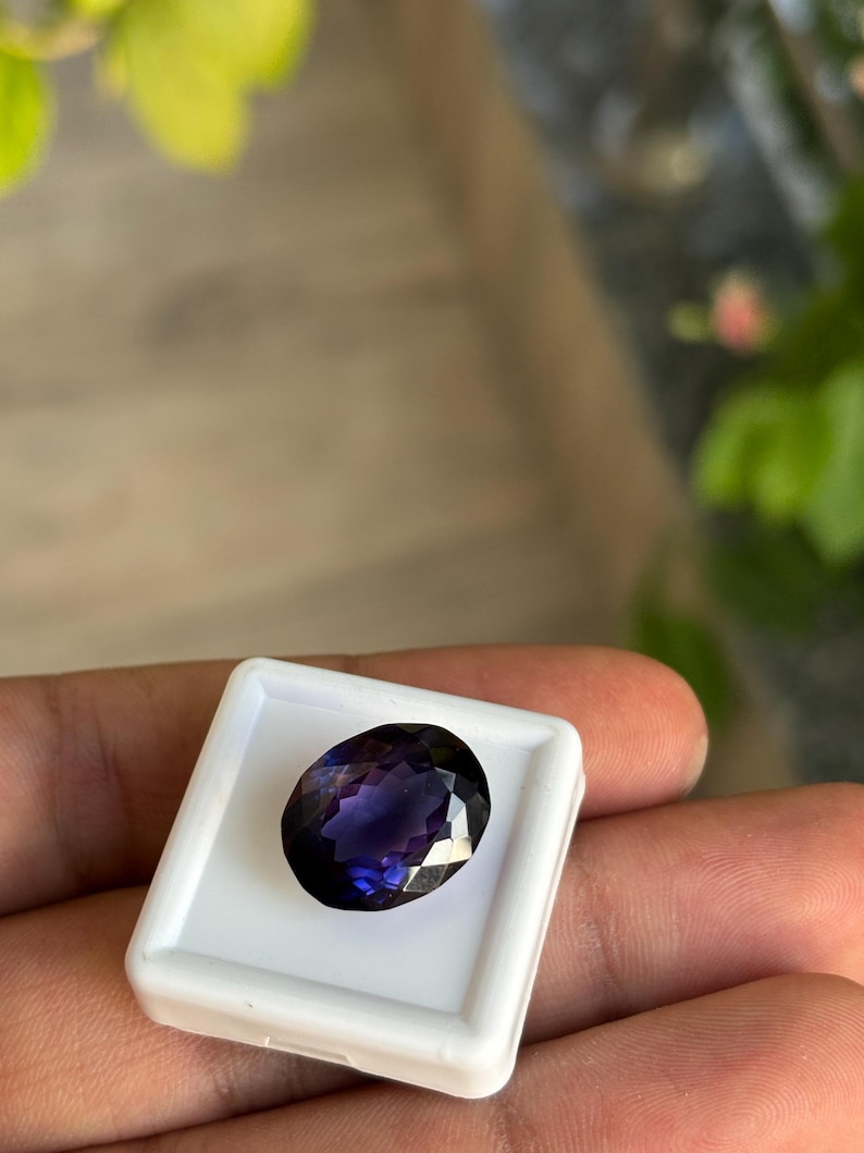May include: A faceted, oval-shaped, deep purple gemstone set in a white square display case. The gemstone has a brilliant cut, reflecting light. The background is blurred, with hints of green foliage and a wooden surface.