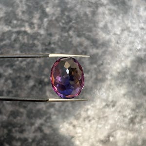 May include: An oval-cut gemstone with a faceted surface, held by tweezers. The gem displays a gradient of colors, transitioning from purple to pink and blue. The gem is approximately 1 inch in length.