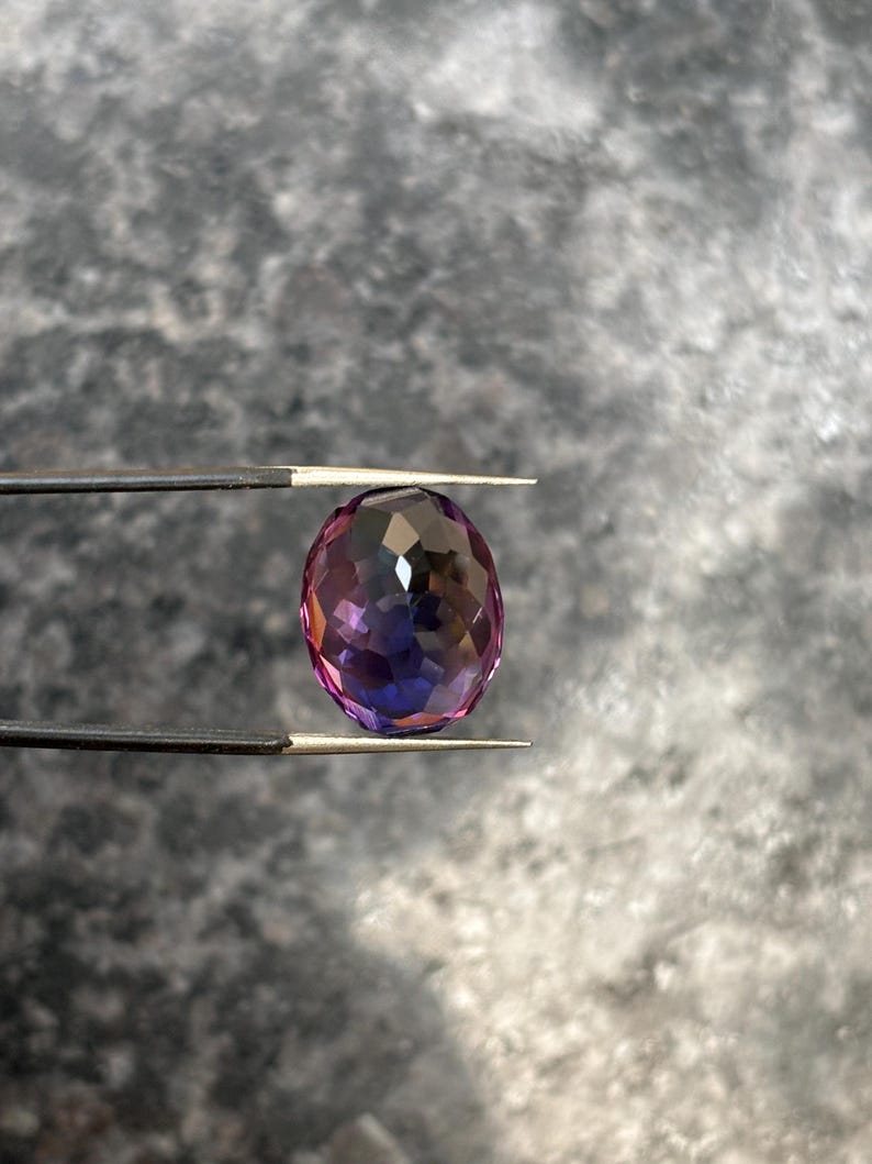 May include: An oval-cut gemstone with a faceted surface, held by tweezers. The gem displays a gradient of colors, transitioning from purple to pink and blue. The gem is approximately 1 inch in length.