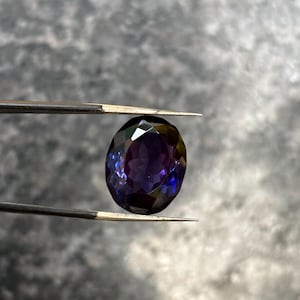 May include: A faceted, oval-shaped gemstone held by tweezers. The gem displays a deep purple hue with flashes of blue and green, reflecting light. The background is a blurred, neutral gray.