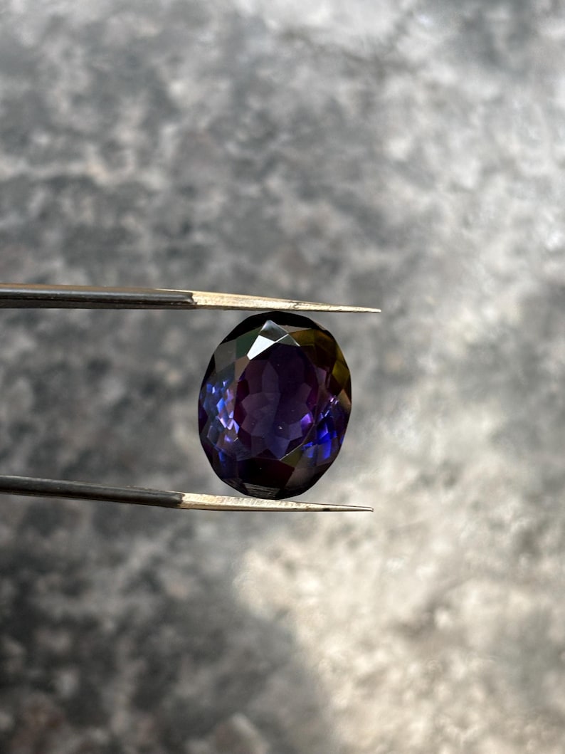 May include: A faceted, oval-shaped gemstone held by tweezers. The gem displays a deep purple hue with flashes of blue and green, reflecting light. The background is a blurred, neutral gray.