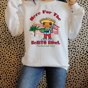 Puede incluir: Sudadera blanca con un gráfico colorido. El gráfico presenta un personaje parecido a un lagarto sosteniendo una taza y una bandera, con el texto "Here For The Benito Bowl" y "The Best Half Time".