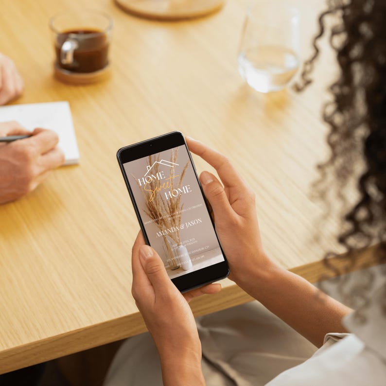 May include: A person holding a black smartphone displaying a "Home Sweet Home" design. The design features a house outline, text, and a vase with dried plants. The background is a soft beige color.