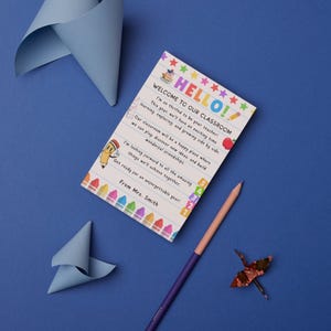 May include: A welcome to the classroom card with the text "HELLO!" and a message from Mrs. Smith. The card is decorated with colorful stars, crayons, and a pencil. A purple and a peach colored pencil, and origami are also on the blue surface.
