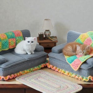 May include: Miniature gray sofas with colorful crocheted blankets and trim. A white cat sits on one sofa, and an orange cat sleeps on the other. A small rug is on the floor, and a lamp and side table are in the background.