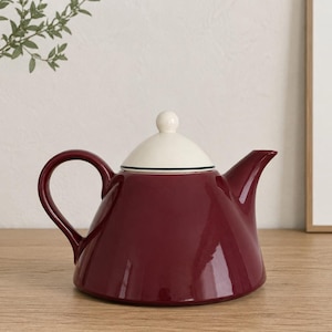 May include: A deep burgundy ceramic teapot with a wide, rounded base, a curved handle, and a pointed spout. The teapot features a removable, smooth cream-coloured lid with a small knob handle and a thin dark rim detail at the base of the lid.