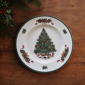 May include: A white ceramic dinner plate featuring a central green Christmas tree decorated with ornaments and gifts. The rim is accented with a dark green border and repeating motifs of red ribbons, holly leaves, and small gold bells.