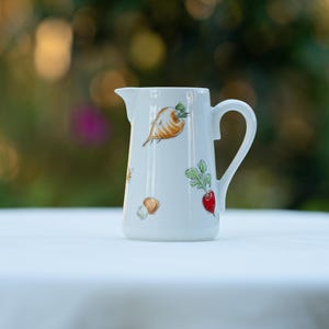 May include: A white ceramic jug featuring a painted pattern of various vegetables including a parsnip, a red radish with green leaves, and a small brown mushroom. The jug has a curved handle and a pouring spout on the upper rim.