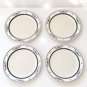 May include: A set of four round ceramic plates with a cream-coloured centre and a wide rim. The rim features a dark blue circular band decorated with repeating floral motifs in pink, green, and blue, interspersed with small red dots.