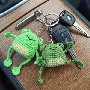 May include: Two handmade crochet frog keychains in shades of green and cream, with black stitched details. The keychains are attached to a set of car keys, resting on a wooden surface.