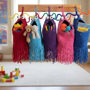 May include: Six colourful, knitted monster-shaped storage bags hang from a wooden rack. Each bag has googly eyes and fringe, and is filled with toys, pencils, and books. The bags are red, blue, purple, pink, and blue.