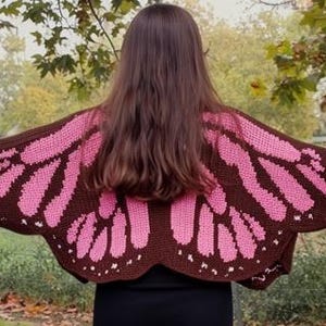 Puede incluir: Un chal de ala de mariposa de ganchillo marrón y rosa. El chal presenta una base marrón con patrones de alas rosas. La persona está de pie afuera con los brazos extendidos, mostrando el diseño del chal. El chal es un accesorio de moda único.