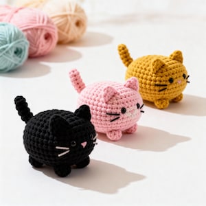 Könnte beinhalten: Drei gehäkelte Katzenfiguren in Schwarz, Rosa und Gelb, jede mit kleinen Ohren, einem Schwanz und gestickten Gesichtszügen. Die Katzen befinden sich auf einer weißen Oberfläche, im Hintergrund Wollknäuel.