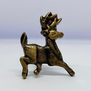 May include: A bronze-coloured figurine of a deer in a running pose. The deer has detailed antlers and a textured surface, giving it a vintage or antique appearance. The figurine is set against a plain white background.
