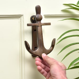 May include: Bronze-colored anchor-shaped door knocker. The nautical-themed knocker is mounted on a cream-colored door. The anchor design features a crossbar, a ring, and curved flukes. The knocker is made of metal and has a rustic finish.