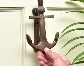 May include: Bronze-colored anchor-shaped door knocker. The nautical-themed knocker is mounted on a cream-colored door. The anchor design features a crossbar, a ring, and curved flukes. The knocker is made of metal and has a rustic finish.