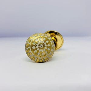 May include: A decorative door knob with a white and gold floral design. The knob is round with a gold base and a detailed pattern. The design features a central flower and intricate details.