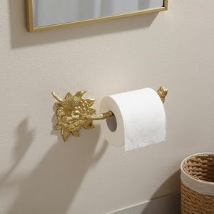 Op de afbeelding: Een goudkleurige, decoratieve toiletpapierhouder met een vogel- en bloemmotief. De houder is aan een lichtgekleurde muur bevestigd en bevat een rol wit toiletpapier. Een spiegel is zichtbaar op de achtergrond.