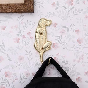 Op de afbeelding: Een goudkleurige metalen wandhaak in de vorm van een hond. De haak is bevestigd op bloemenbehang en houdt een zwarte tas vast. De hond zit met zijn hoofd opzij. De haak is bevestigd met drie schroeven.