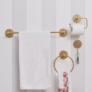Könnte beinhalten: Ein Set goldfarbener Badezimmerarmaturen, darunter eine Handtuchstange mit einem weißen Handtuch, ein Toilettenpapierhalter, ein Haken mit Schlüsseln und ein Handtuchring mit einem rot-weiß gemusterten Handtuch. Die Armaturen haben verzierte Details.