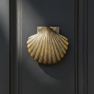 以下が含まれることがあります： 暗い灰色のドアに取り付けられた真鍮製の貝殻型のドアノッカー。ノッカーには詳細な溝があり、長方形の取り付けプレートが付いています。ドアには垂直パネルがあります。
