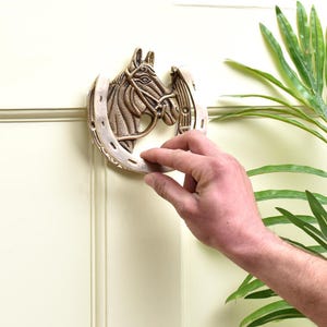 May include: A bronze-colored horse head door knocker with a horseshoe design. The horse head features detailed bridle and mane elements. The knocker is mounted on a cream-colored door, with a hand reaching for the knocker.