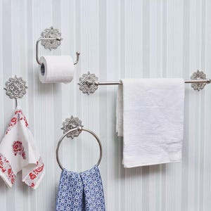 May include: A set of ornate, silver-toned bathroom fixtures, including a towel bar, toilet paper holder, towel ring, and two hooks. The fixtures have a decorative floral design. A white towel and patterned hand towels are displayed.
