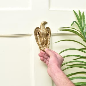 May include: A brass eagle door knocker, mounted on a white door. The eagle is detailed with feathers and a prominent beak. The knocker is held by a hand. Decorative hardware for home entryways.