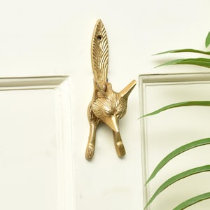May include: A gold-coloured fox-shaped hook. The metal hook features a detailed design of a fox's head, tail, and legs. It is mounted on a white door. The fox's tail extends upwards, and the hook is formed by the fox's legs. A decorative item.