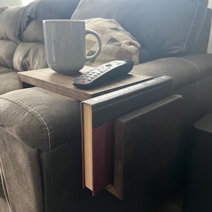 May include: A gray mug, remote control, and a wooden tray rest on the arm of a brown sofa. A book holder is attached to the sofa arm, holding two books. The scene suggests a cozy living room setting.