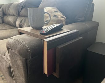 May include: A gray mug, remote control, and a wooden tray rest on the arm of a brown sofa. A book holder is attached to the sofa arm, holding two books. The scene suggests a cozy living room setting.