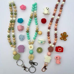 Op de afbeelding: Drie kralen lanyards in groen, teal en bruin, met diverse bedels. Bedels zijn onder meer bloemen, een spook, een peperkoekmannetje en medische items. Elke lanyard heeft een metalen sluiting.