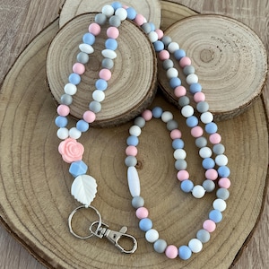 Peut inclure: Un cordon composé de perles en silicone rondes dans des tons bleu clair, rose tendre, blanc et gris. Le design comprend une perle en forme de rose rose, une perle hexagonale bleu clair et une perle en forme de feuille blanche avec un mousqueton.