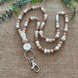 May include: A beaded lanyard in shades of brown, beige, and white. The lanyard features a daisy charm and a silver-colored clasp. The beads are various sizes and shapes, creating a decorative accessory.