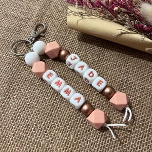 Puede incluir: Dos llaveros personalizados con los nombres "EMMA" y "JADE" deletreados con cuentas de letras blancas. Los llaveros presentan cuentas geométricas de color melocotón, cuentas de color bronce y un cierre plateado. El fondo es una superficie texturizada y marrón claro.