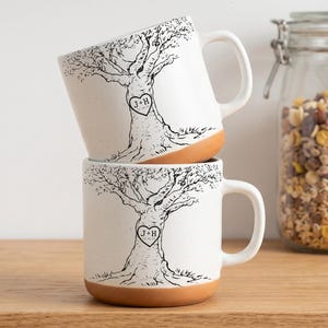 Peut inclure: Deux tasses en céramique empilées verticalement, avec une finition blanche mouchetée et une base en terre cuite. Chaque tasse présente une illustration au trait noir d'un arbre avec un cœur gravé dans le tronc contenant J+H et une date.