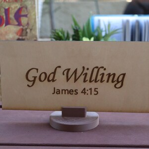 Può includere: Un cartello in legno con le parole "God Willing" e "James 4:15" incise in un carattere decorativo. Il cartello è esposto su una piccola base rotonda di legno. Lo sfondo include un libro e del verde.