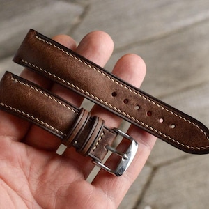 Puede incluir: Una correa de reloj de cuero marrón con costuras blancas y una hebilla plateada. La correa se sostiene en una mano, mostrando su detallada artesanía y diseño clásico. Un accesorio elegante.