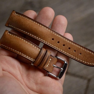 Op de afbeelding: Een paar lichtbruine leren horlogebandjes met witte stiksels en een zilveren gesp. De bandjes worden in een hand gehouden, waarbij de gedetailleerde stiksels en de kwaliteit van het leer worden getoond. De horlogebandjes zijn nieuw.
