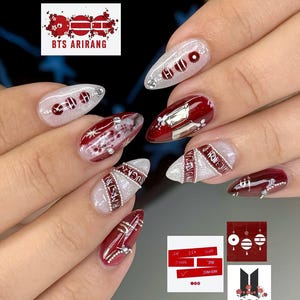 May include: A set of manicured nails with a design featuring red and white accents, silver embellishments, and the text "BTS ARIRANG". The nails have a glossy finish and include various nail art elements.