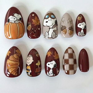 May include: A set of ten oval false nails with autumn-themed designs. The nails feature Snoopy, Charlie Brown, acorns, and fall leaves. Colours include brown, orange, and beige. One nail has a croissant design, and another has a brown and beige checkered pattern.