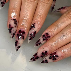 May include: Close-up of long, pointed fingernails with a nude base and dark burgundy floral designs. The nails feature intricate, hand-painted flowers with silver accents, creating a sophisticated and artistic look.
