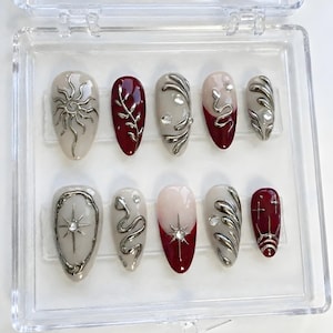May include: A set of ten artificial nails in a clear plastic case. The nails feature a variety of designs, including silver sunbursts, leaves, and swirls, with a color palette of beige, burgundy, and silver. Some nails have a gradient design.