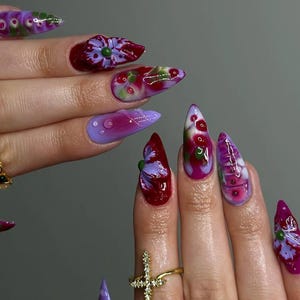 Könnte beinhalten: Ein Set aus langen, spitz zulaufenden Acrylnägeln mit aufwendigen Blumendesigns. Die Nägel weisen eine Vielzahl von Farben auf, darunter Lila, Rot, Grün und Weiß, mit detaillierten Blumen- und Wassertropfen-Akzenten. Goldringe schmücken die Finger.