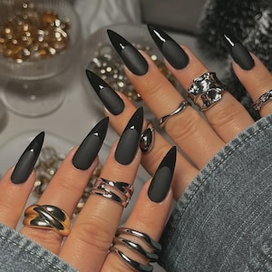 May include: Long, matte black stiletto nails with glossy black tips. The hand is adorned with various silver and gold rings. The nails are a bold fashion statement.
